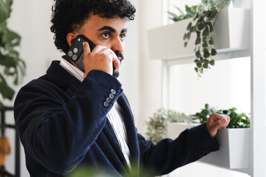 A man in a suit talks on his phone while standing near a window with plants.