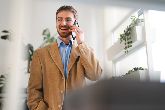 A smiling man in a corduroy jacket talks on his mobile phone in a modern office setting with plants.