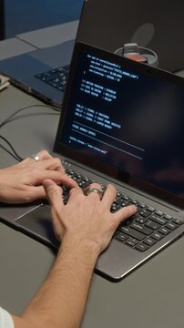 Vertical shot of hands of unrecognisable developer wearing silver rings coding on laptop at work table in dim lighting