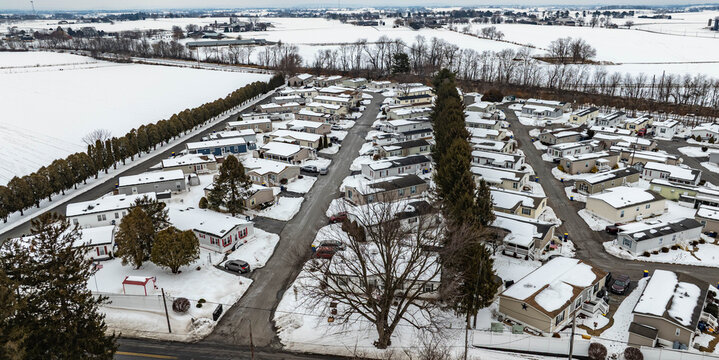 A mobile home park is seen covered in snow during winter. The scene shows rows of homes surrounded by white snow and a few trees. Fields stretch out in the background under a gray sky.