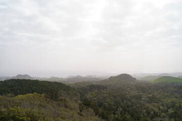 Rolling green hills of the Azores, Portugal, stretch into soft haze in the distance. The landscape shows layered terrain, rural fields, and Atlantic island scenery under a calm sky.