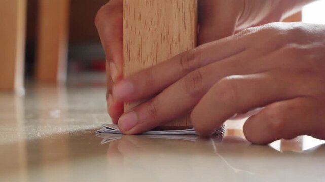 A person puts folded paper under a dining table leg to stabilize the table