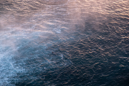 An abstract top-down perspective of the Mediterranean Sea surface, featuring soft sea spray and mist illuminated by the warm golden light of sunrise. The deep blue water creates a beautiful contrast w