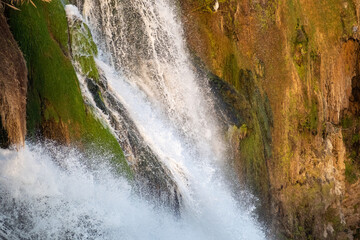 A detailed close-up photograph capturing the powerful flow of the Lower Duden Waterfall as it cascades over rugged, brown rocky cliffs. The image highlights the vibrant green moss covering the wet roc © osmanmaasoglu