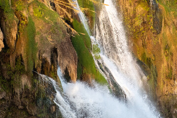 A detailed close-up photograph capturing the powerful flow of the Lower Duden Waterfall as it cascades over rugged, brown rocky cliffs. The image highlights the vibrant green moss covering the wet roc © osmanmaasoglu