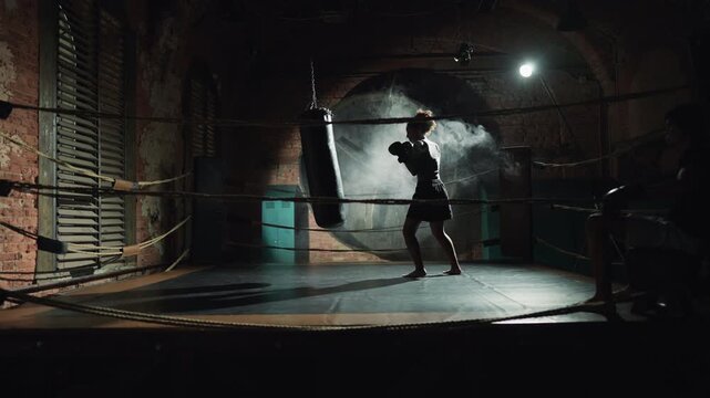 Young woman fighting with punching bag in boxing ring, professional athlete. Healthy lifestyle and sport activity, motivated sportswoman training, physical exercises for release aggression and tension