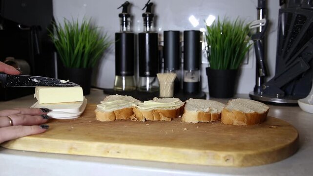 Human hands spreading creamy yellow butter on white baguette bread slices on wooden cutting board. Modern kitchen interior with plants and dispensers in background.