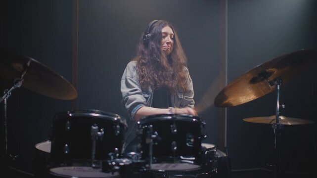 Beautiful drummer girl with long hair is tapping rhythm and drum part in rehearsal room. Young female with headphones performs solo on stage, kit. Sound checking equipment before a rock concert