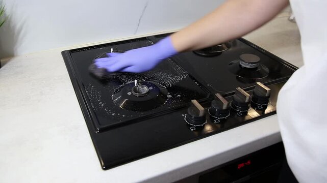 Close up detail view of person hand wearing blue latex glove scrubbing black gas stove burners with soapy sponge to remove dirt. Cleanliness and hygiene of kitchen appliance. Domestic routine for mode
