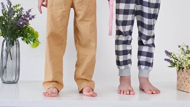 Two children no face standing barefoot on white table, wearing casual home pants, cozy lifestyle. Simple Living, Scandinavian Inspired. Wiggle toes and do foot exercises to combat flat feet