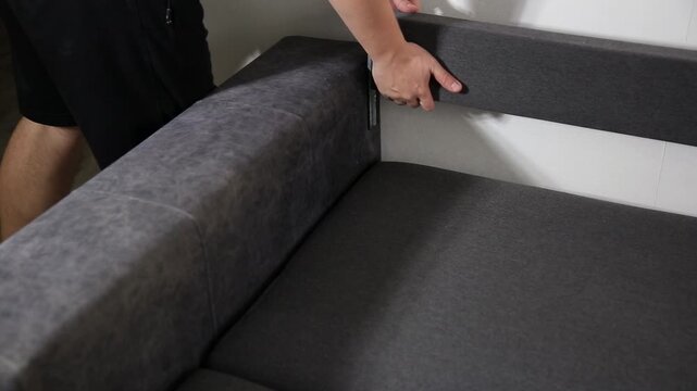 Hands of person carefully fitting backrest section of modern grey sofa against white wall. Home furniture assembly demonstration showing detail of connection.