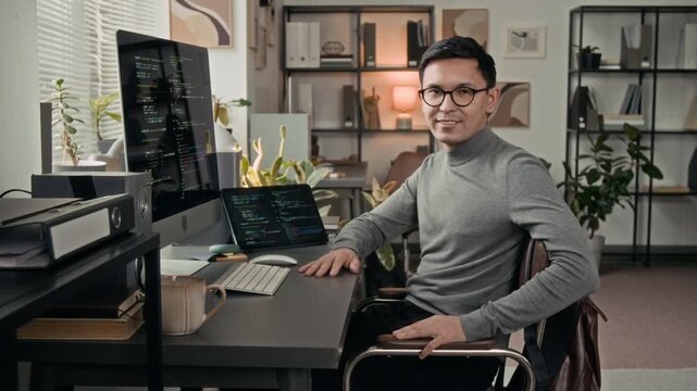 Portrait of positive Caucasian young male software engineer wearing grey roll-neck shirt sitting at work table featuring computer monitor, wireless keyboard, digital tablet and mug in IT office