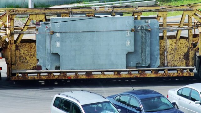 Heavy transport truck with rusty flatbed trailer moves large gray concrete slabs. Industrial cargo for construction project. Parked cars below show scale.