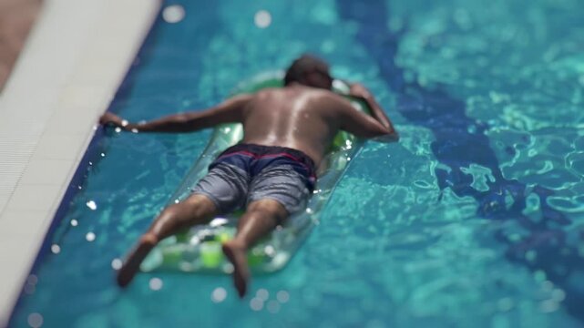 Overhead view shows person on inflatable air mattress, floating in blue swimming pool. Person wears swim shorts. Water reflects sunlight, bright bokeh. Scene conveys peaceful summer relaxation. Slow m