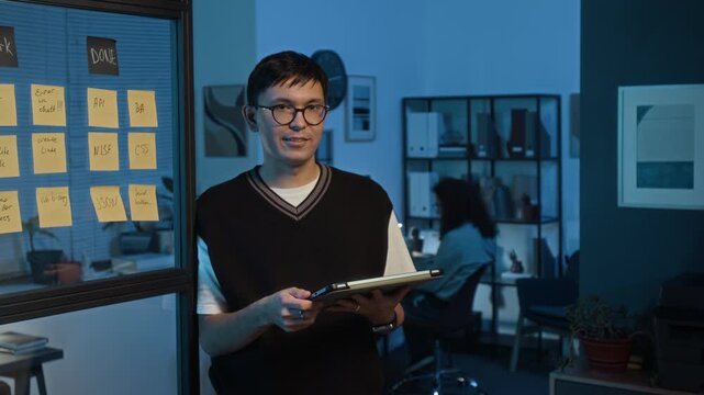 Waist-up portrait of Caucasian young product manager wearing white t-shirt and black vest using digital tablet and looking at camera during workday in dark IT office