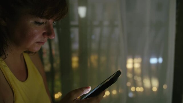 Female Standing Near Window at Night Using Smartphone for Reading News and Messages with Windy City View