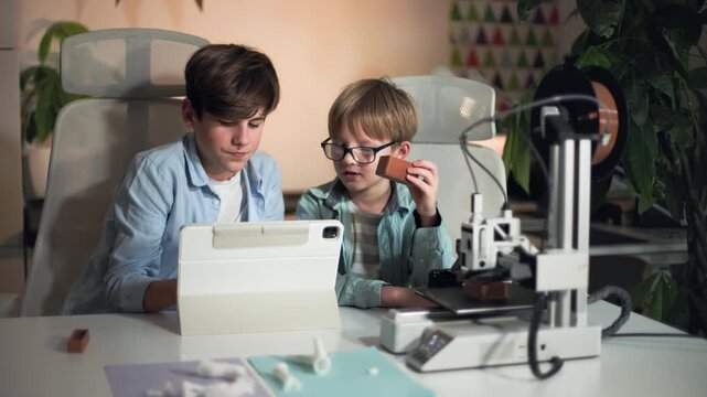 At a white bright table, two kids control 3D printer and discuss printed part