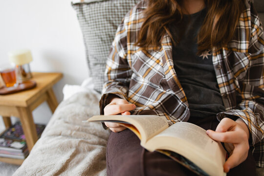 Person sitting on sofa reading a book in comfortable home setting. Soft blanket and warm natural light create peaceful mood. Concept of cozy lifestyle, leisure time, education and mindful rest.