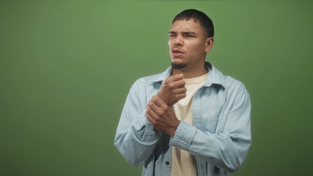 Young hispanic man clutching wrist and wincing, holding the painful joint while wearing a light denim shirt on a green studio set; pain injury recovery.
