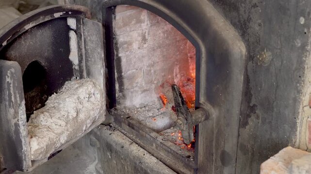 Close-up of an African baker opening a wood-fired brick oven and adding logs to glowing embers. Traditional heat source used for artisan baking and rustic food preparation.
