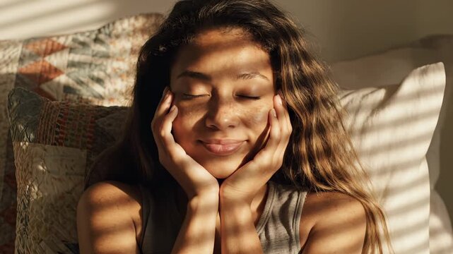 Woman relaxes on sofa with sunlight shadows dancing on face. Female resting peacefully with hands on cheeks. Calm woman enjoys sunlight at home. Relaxed female on sofa with shadow patterns on face.