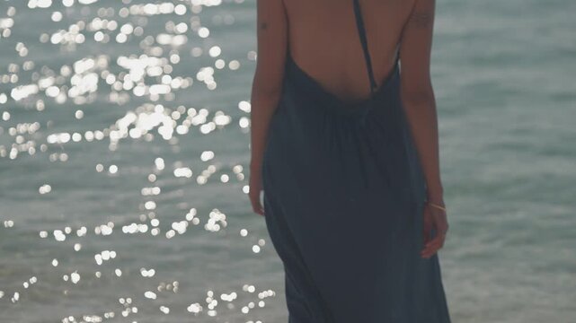 Woman in backless dress by sunlit sea, exotic luxurious lifestyle shot in slow motion walking toward the sea