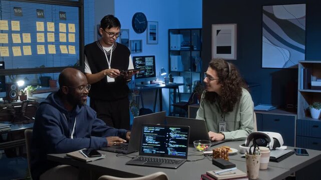 Young group of three diverse male IT specialists using laptops and tablet while discussing software development in modern office illuminated by desk lamps