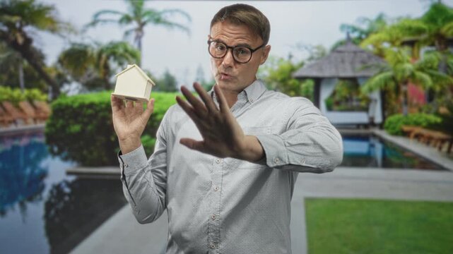 Man holding small house model with palm out stop gesture and glasses in building poolside garden; caution negotiation.