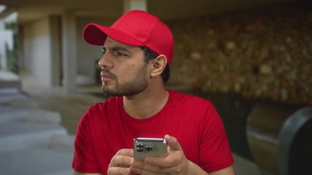 Man in red cap frowns and holds smartphone with both hands while leaning forward slightly in spa building; uncertainty.