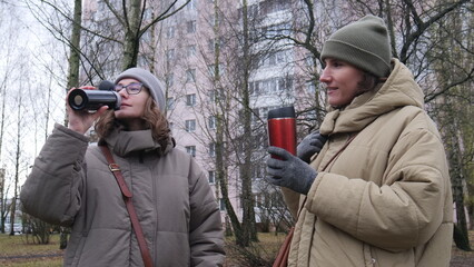 Two young women in park parkas and beanies warming up with reusable thermo cups on a cold morning, chatting and smiling during a cozy urban break together © Aliaksandr