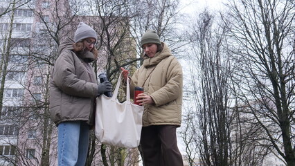 Naklejka premium Two young women friends standing outdoors in winter clothing, putting personal thermoses into or taking them out of a reusable fabric tote bag, sharing a warm drink