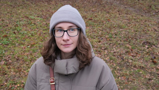 Young woman confidently looking away, wearing a beanie and glasses, standing outdoors in a park enjoying a casual autumn day with dry leaves and grass on the ground