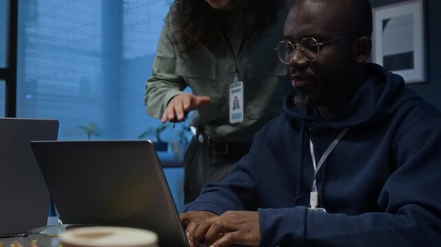 Black male developer with glasses coding on laptop and listening to advice of long-haired Caucasian guy standing beside his desk