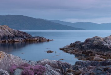 Obraz premium Coastal landscape featuring rocky shoreline, serene water, distant mountains under a dusky sky