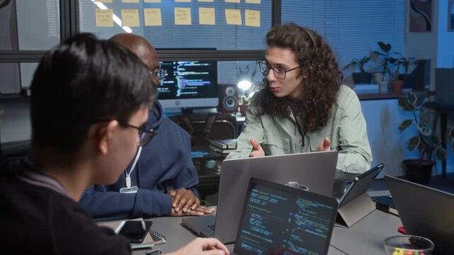 Diverse team of young programmers designing software in dimly lit IT office, two of them conversing pointing at laptop screen, third one writing code in foreground