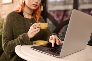 Charming woman with red hair enjoys coffee while working on laptop in modern cafe
