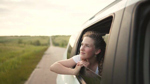 Young woman leans from car window looking at winding road and green countryside field smile and daydreaming while sunlight warms face hair blowing in breeze travel and journey feeling freedom