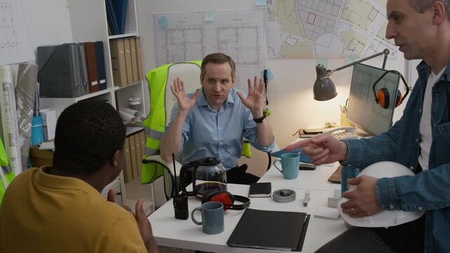 Zoom out shot of three diverse engineers arguing while discussing and planning work at desk in small construction office with blueprints