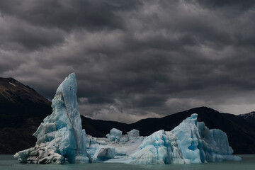 Electric blue iceberg floating in a glacial lake under heavy, dark storm clouds with silhouetted mountains in the background. High contrast moody nature photography. © marcodenaro