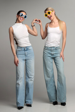 Full length studio shot of two young beautiful women best friends wearing funny party glasses making heart shape gesture in white tops and jeans on gray background