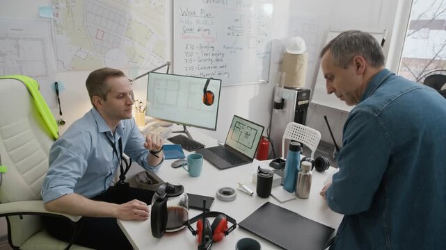 Handheld shot of adult foreman giving documents to engineer sitting at table with blueprints on computers in small construction office