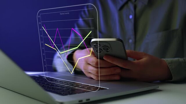 Person checks data trends on a smartphone while using a laptop in a dark room.