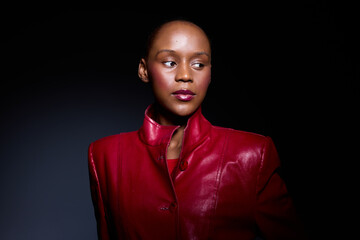 Obraz premium Portrait of a confident dark skinned African woman wearing a red leather jacket, looking sideways against a dark background. Concept of fashion, elegance, and empowerment.