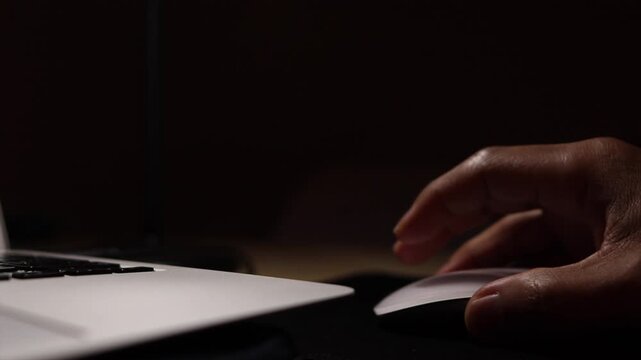 Person's hand clicking and scrolling on a computer mouse next to a modern laptop in a dark office room at night