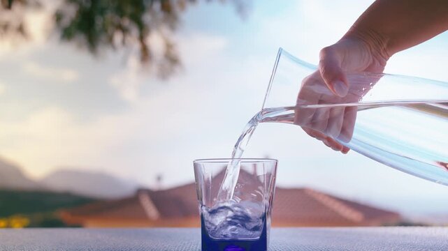 Female Hand Pouring Clear Water from Pitcher into Glass Outdoors Under Blue Sky