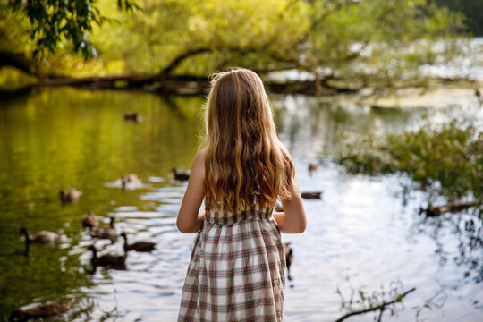 Little elementary school girl feeding wild ducks on a pond. Cute child enjoying nature and animals outdoors in summer, happy kid with eye glasses spending time near water.