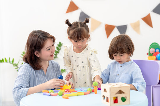 Asian single mother sits beside round table, guiding toddler girl, preschool boy sorting wooden toys, learning colors shapes counting, practicing coordination, teamwork, warm family bonding indoors.