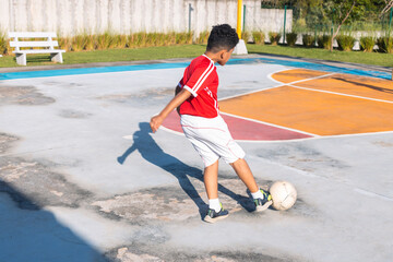 Obraz premium A young boy in a red and white shirt kicks a soccer ball on a concrete court