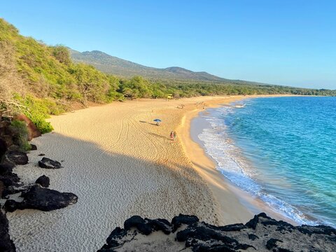 Golden sands stretch along Big Beach in Maui near sundown, where gentle waves meet turquoise waters beneath lush hills and clear skies.