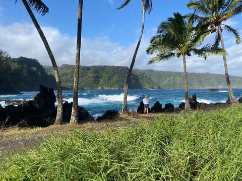 Senior man admiring a dramatic coastal view along Maui&rsquo;s Road to Hana, framed by palm trees, lava rocks, waves, and lush tropical cliffs.